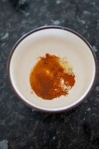 A small bowl brimming with a mix of ground spices, mainly red and orange, rests on a dark countertop. Nearby, a scattering of green banana chips adds an unexpected touch of color and crunch.