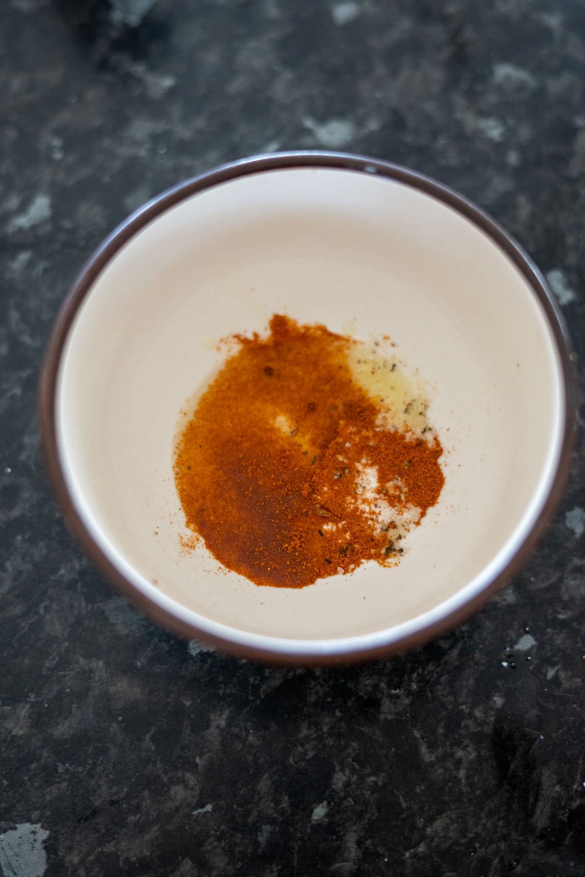 A small bowl brimming with a mix of ground spices, mainly red and orange, rests on a dark countertop. Nearby, a scattering of green banana chips adds an unexpected touch of color and crunch.