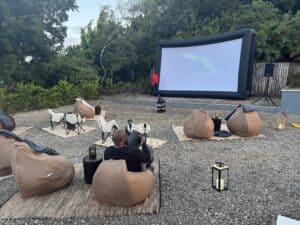 A St. Lucia cinema under the stars, featuring an inflatable screen surrounded by cozy bean bags and chairs. Decorative lanterns add charm to the gravel setup, while a lone figure stands near the screen, enveloped by lush trees and greenery.