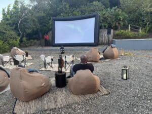 Experience the St Lucia cinema under the stars with an outdoor setup featuring an inflatable screen, cozy bean bags, and rugs on gravel. A few moviegoers relax amidst lush greenery, illuminated by a nearby lantern's warm glow.