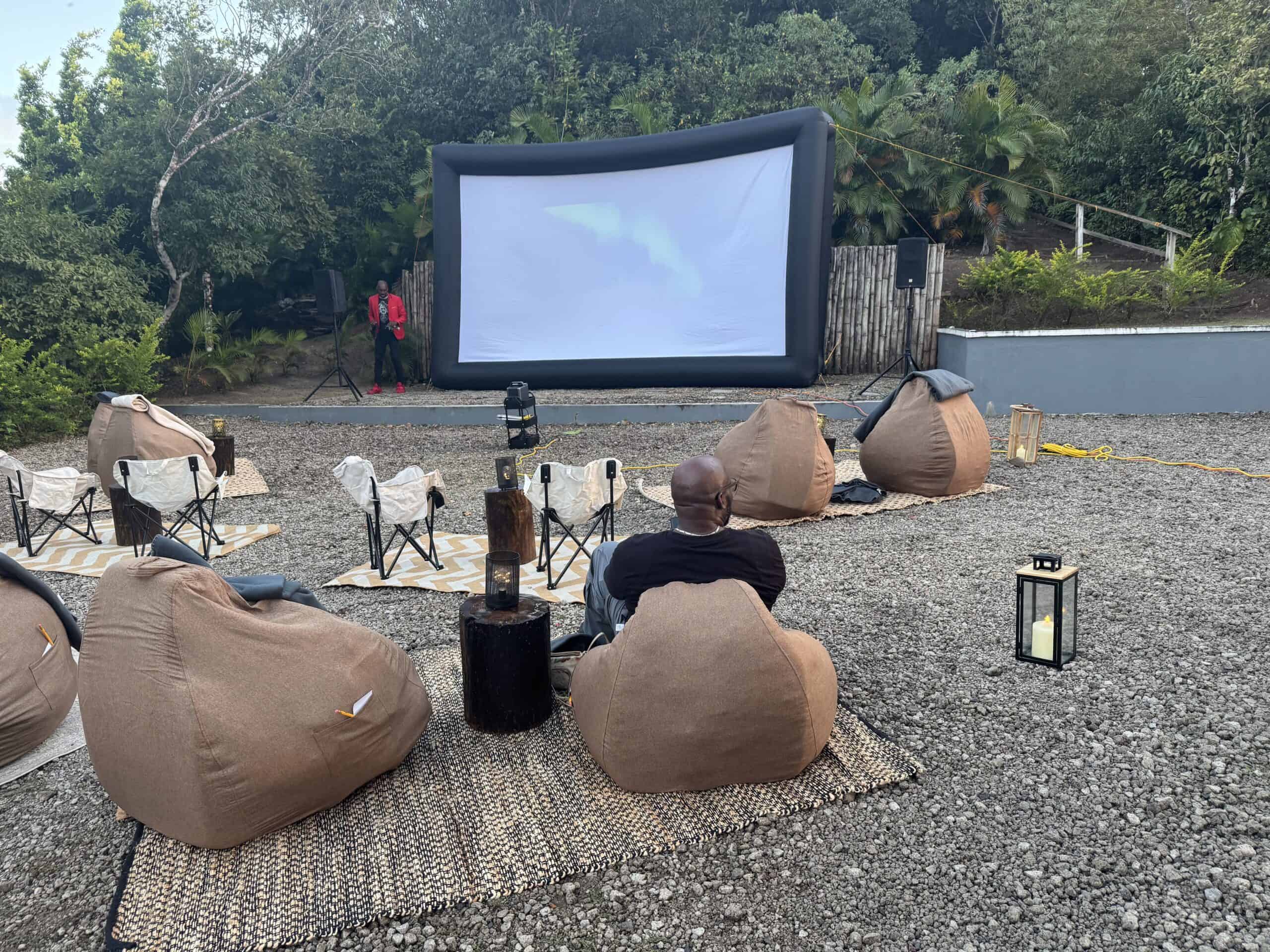 Experience the St Lucia cinema under the stars with an outdoor setup featuring an inflatable screen, cozy bean bags, and rugs on gravel. A few moviegoers relax amidst lush greenery, illuminated by a nearby lantern's warm glow.