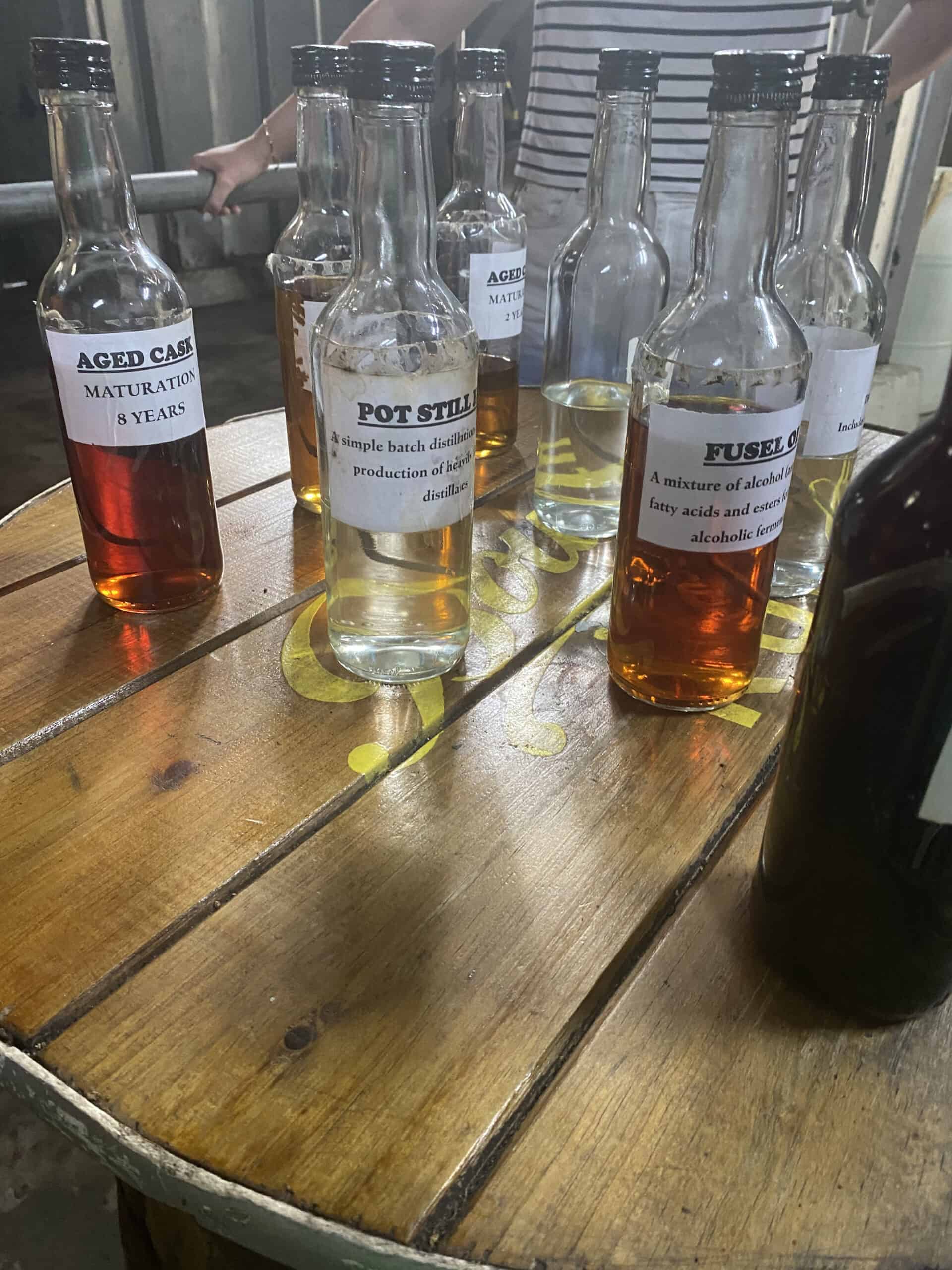 Several labeled bottles, showcasing different stages and components of Saint Lucia rum, are arranged on a round wooden table. A person stands in the background.