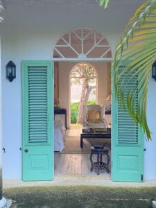 Open mint green double doors reveal a cozy living space—perfect for relaxing after exploring house hunting tips in Saint Lucia—with a white armchair, small table, and a view of lush greenery through an arched window.