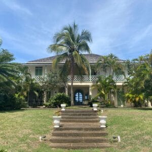 Large, two-story colonial-style house with a balcony, surrounded by palm trees and greenery, viewed from stone steps under a blue sky—perfect for those seeking house hunting tips in Saint Lucia.