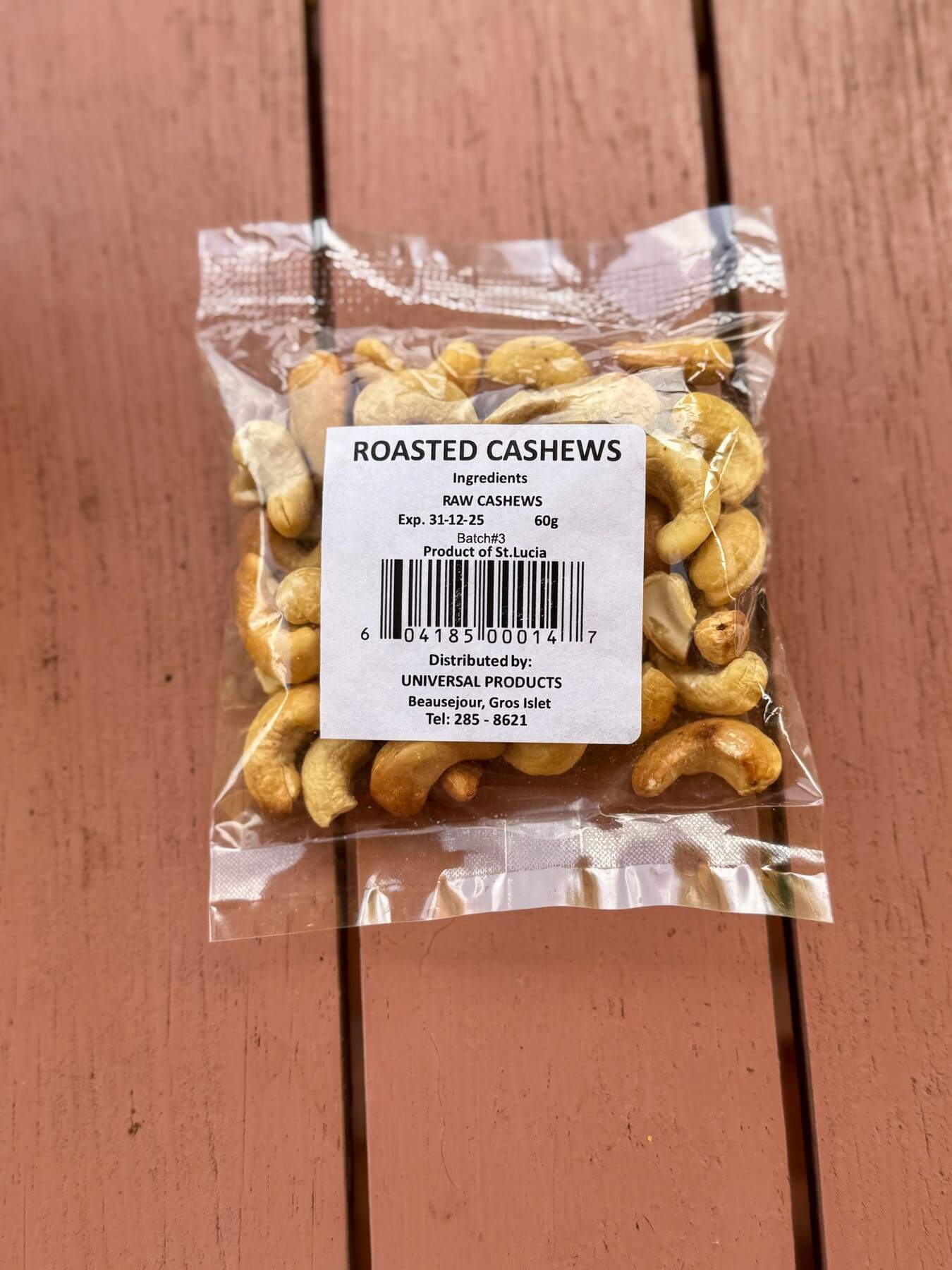 A sealed plastic package of roasted cashews in Saint Lucia, featuring a white label with ingredients, expiration date, weight, and distributor details, rests on a wooden surface.