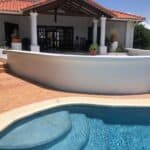 A swimming pool with curved steps sits in front of a white house with a tiled roof and columns, potted plants on the patio—perfect inspiration if you're seeking house hunting tips in Saint Lucia.