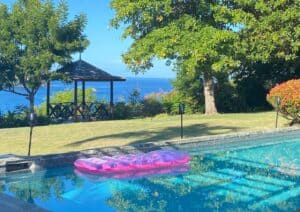 A pink inflatable pool float sits in a clear swimming pool with a garden, gazebo, and stunning ocean view—perfect inspiration for house hunting tips in Saint Lucia on a sunny day.