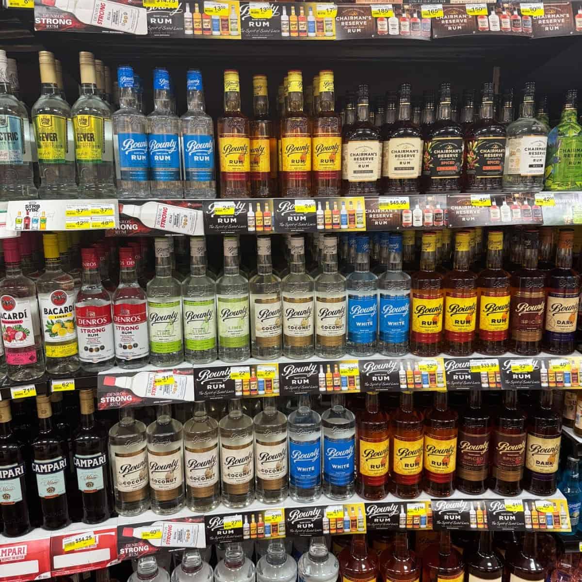 Shelves in a store display various brands and types of rum, including light, dark, and flavored options such as Saint Lucia rum, with price tags visible below each bottle.