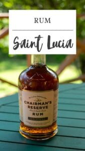 A bottle of Chairman’s Reserve Original Saint Lucia Rum sits on a green table outdoors, with trees and a wooden railing in the background.