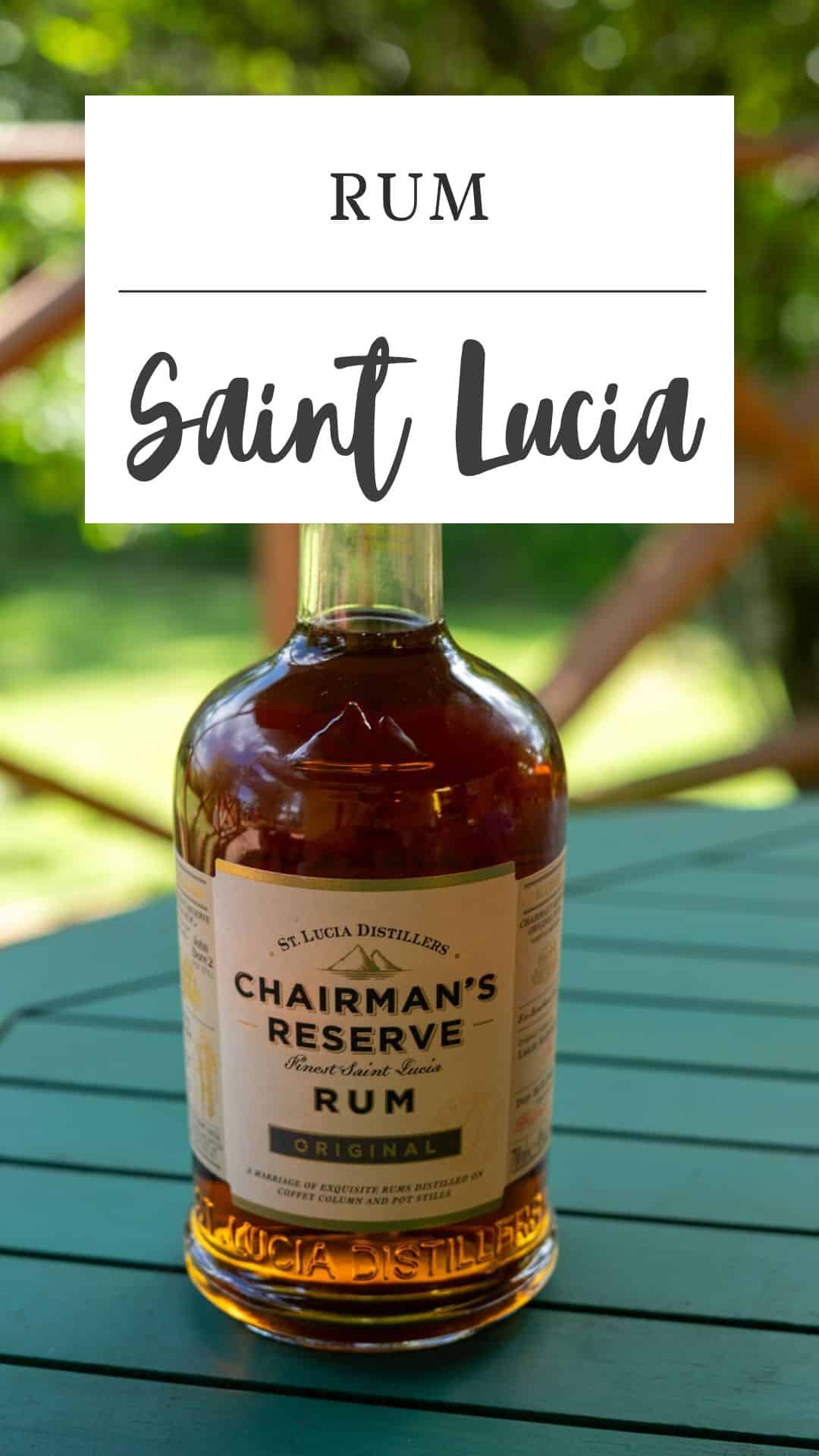 A bottle of Chairman’s Reserve Original Saint Lucia Rum sits on a green table outdoors, with trees and a wooden railing in the background.