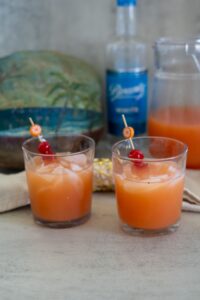 Two glasses filled with an orange-colored drink made with Saint Lucia Rum, ice cubes, and garnished with cherries and orange slices, with a bottle and pitcher in the background.