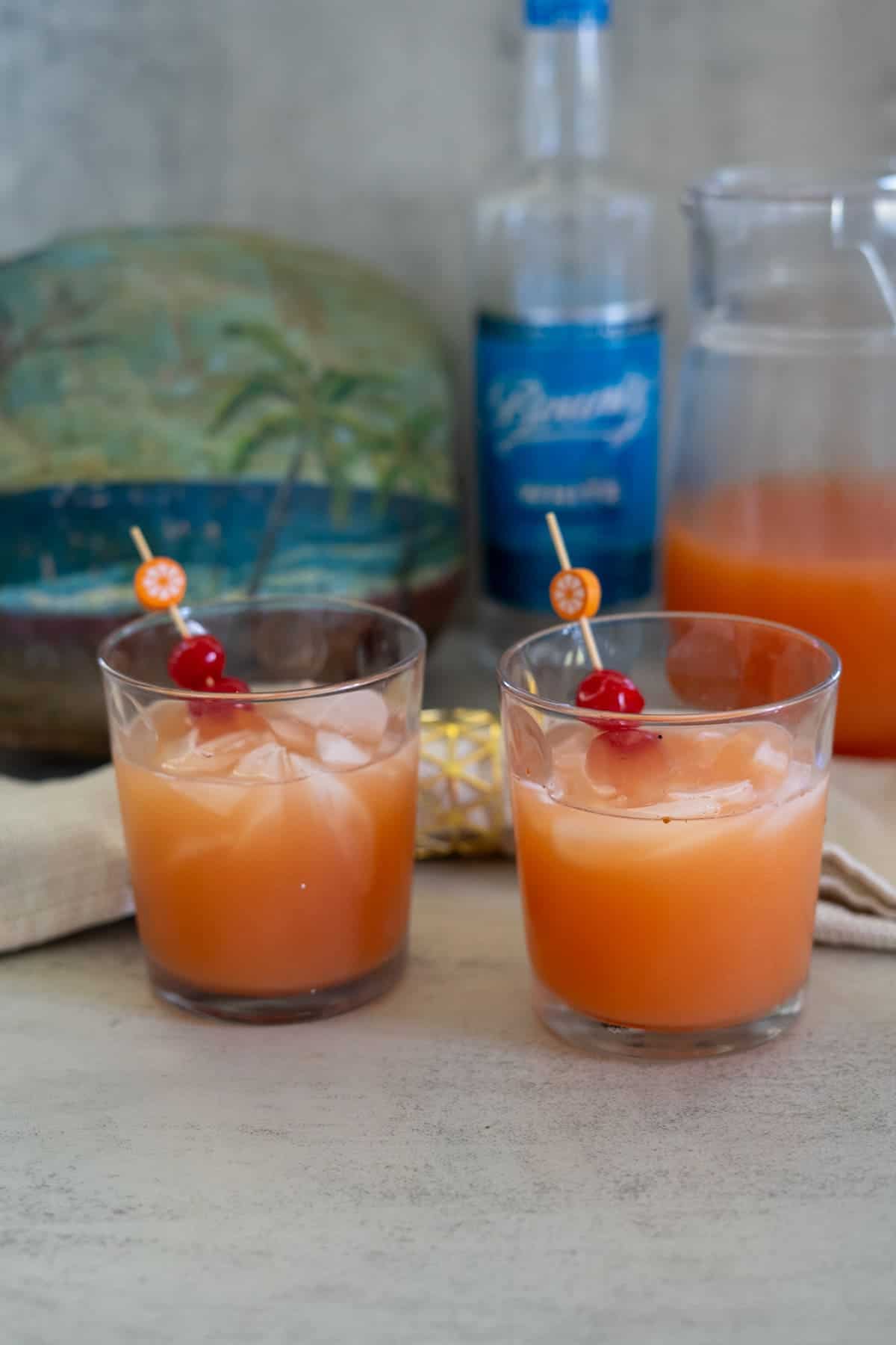 Two glasses filled with an orange-colored drink made with Saint Lucia Rum, ice cubes, and garnished with cherries and orange slices, with a bottle and pitcher in the background.