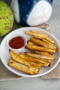 A white plate with baked breadfruit fries and a side of ketchup, placed on a beige napkin. A green breadfruit and a blue and white ceramic container are in the background.