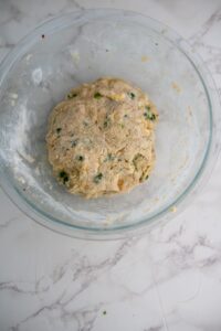 A glass bowl containing a ball of dough with green herbs mixed in, placed on a white marble surface.