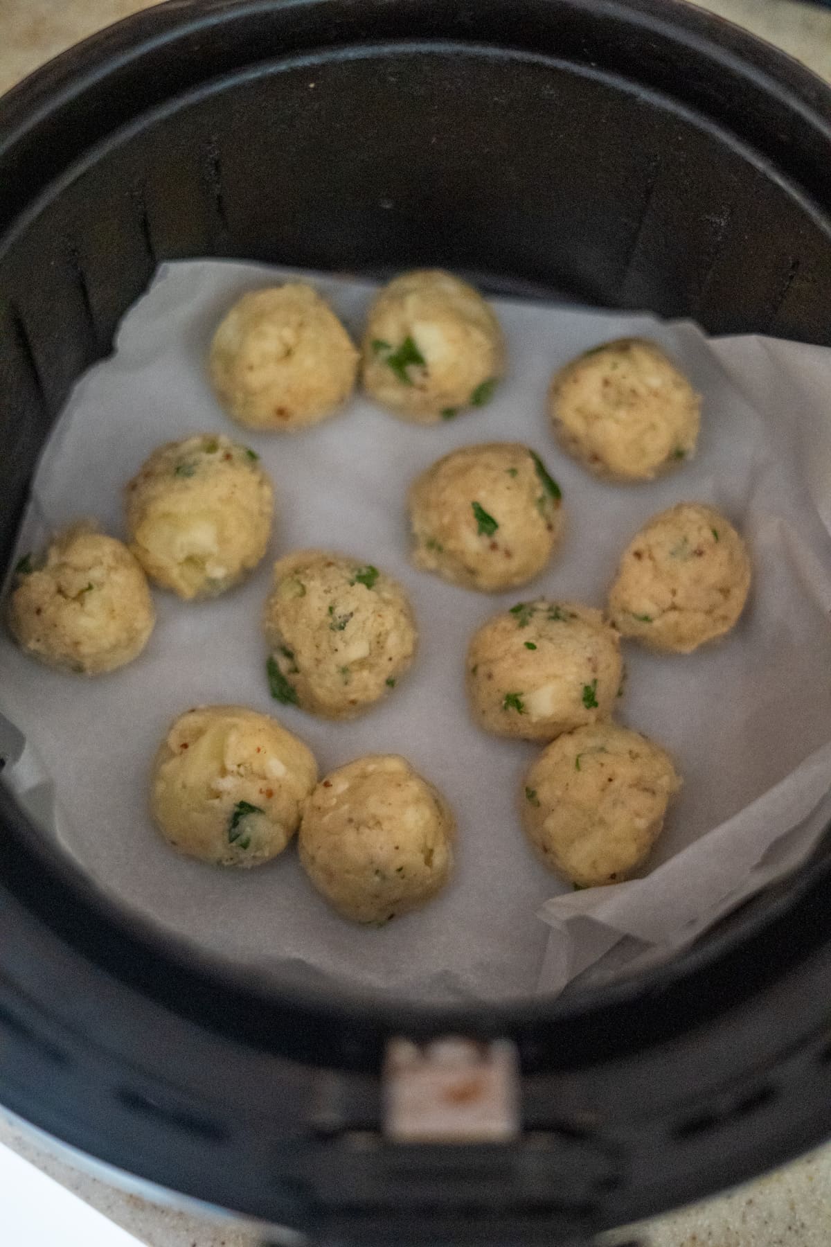 Eleven uncooked, herb-seasoned balls are arranged on parchment paper inside an air fryer basket.