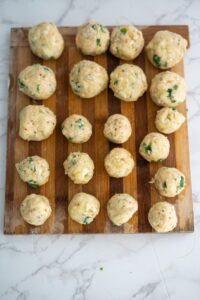 Several uncooked dumpling balls made with herbs and diced ingredients are arranged on a wooden cutting board on a marble surface.