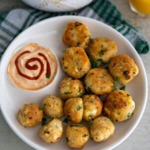 A white plate with a serving of golden-brown veggie balls, garnished with herbs, and a side of dipping sauce decorated with a swirl of sriracha.