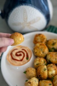 A hand holds a cheese ball above a plate of cheese balls with a swirl of sauce for dipping. A blue and white decorative item is in the background.