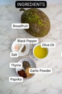 A marble countertop displays labeled ingredients for breadfruit fries: whole breadfruit, olive oil, salt, black pepper, thyme, garlic powder, and paprika in small bowls.