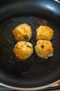 Four golden-brown meatballs are frying in a black nonstick pan with a small amount of oil.