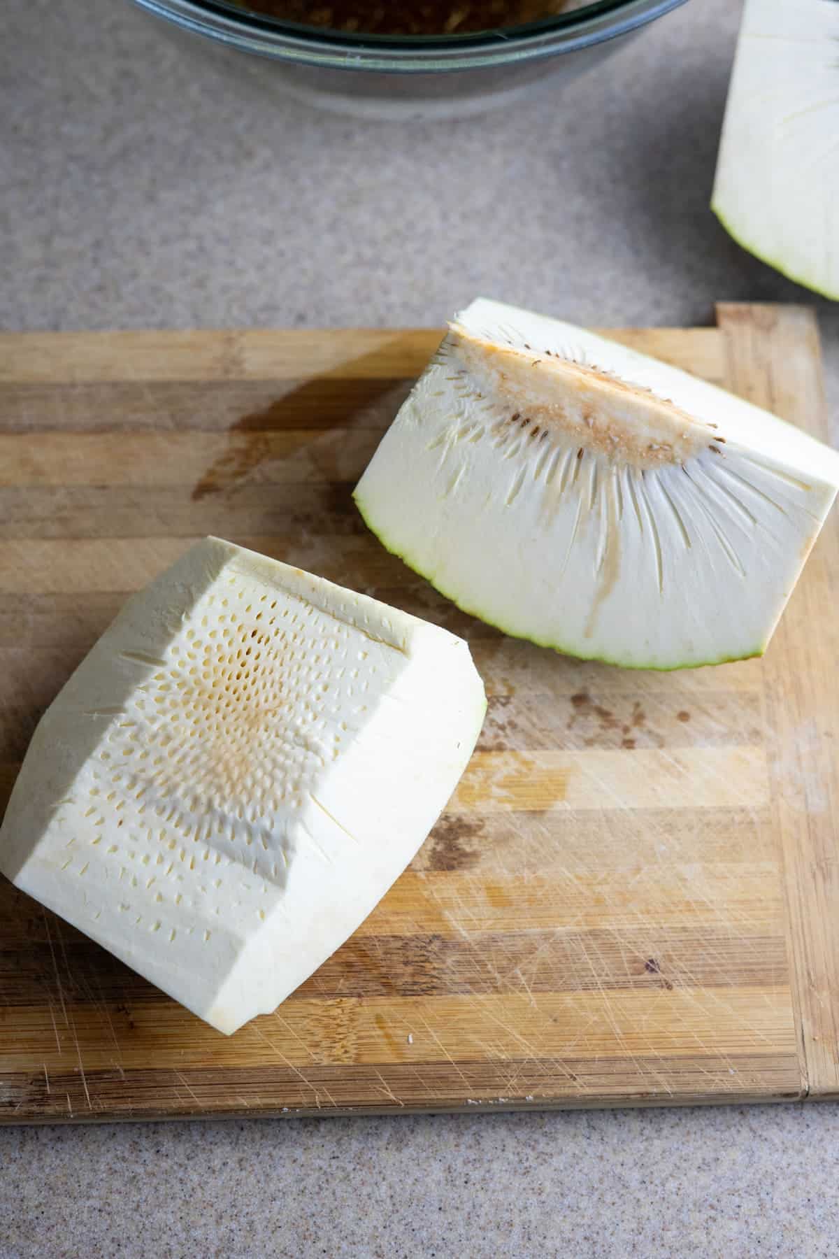 Two pieces of peeled and cut breadfruit sit on a wooden cutting board, ready to be sliced into breadfruit fries, their white flesh and fibrous core clearly visible.