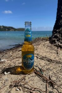 A bottle of Piton Beer stands upright on sandy ground near the shore, with the sea and blue sky in the background.
