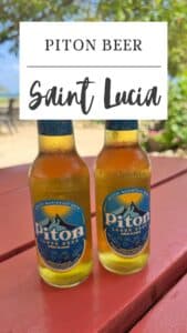 Two bottles of Piton Beer on a red outdoor table, surrounded by lush greenery and a sandy area in the background.