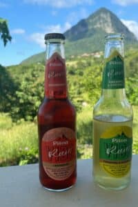 Two bottles of Piton Kube drinks, cranberry and pineapple & coconut flavors, are placed on a ledge with a green mountain in the blurred background, capturing the refreshing spirit of Piton beer.