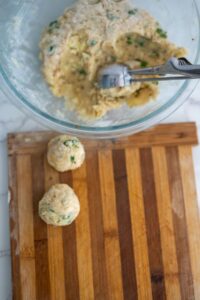 A glass bowl with a mixture and a scoop inside, next to two formed dough balls on a wooden cutting board.
