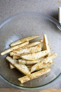 Glass bowl containing seasoned raw breadfruit fries slices on a countertop.