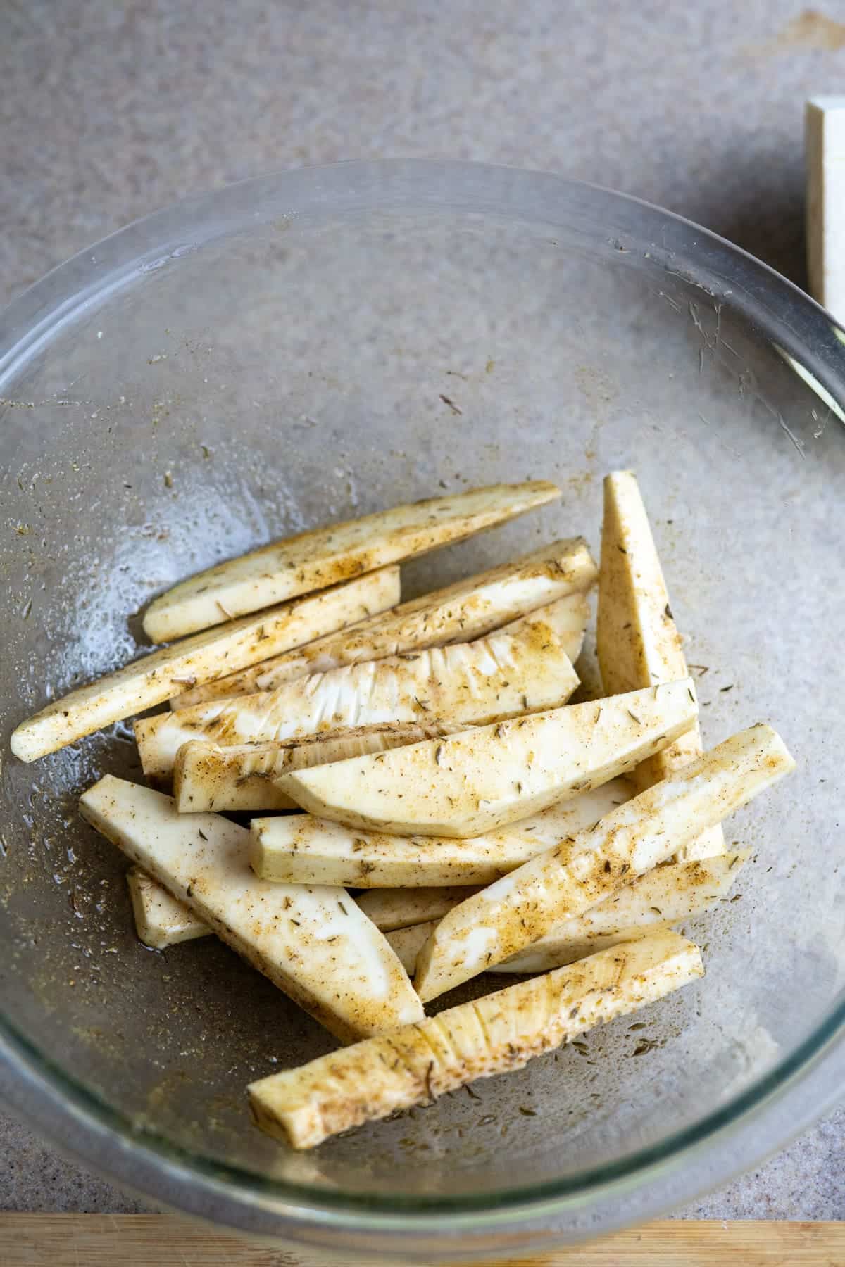 Glass bowl containing seasoned raw breadfruit fries slices on a countertop.