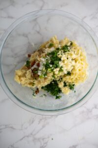 A glass bowl with grated cheese, chopped onions, herbs, spices, and mashed potatoes on a marble surface.