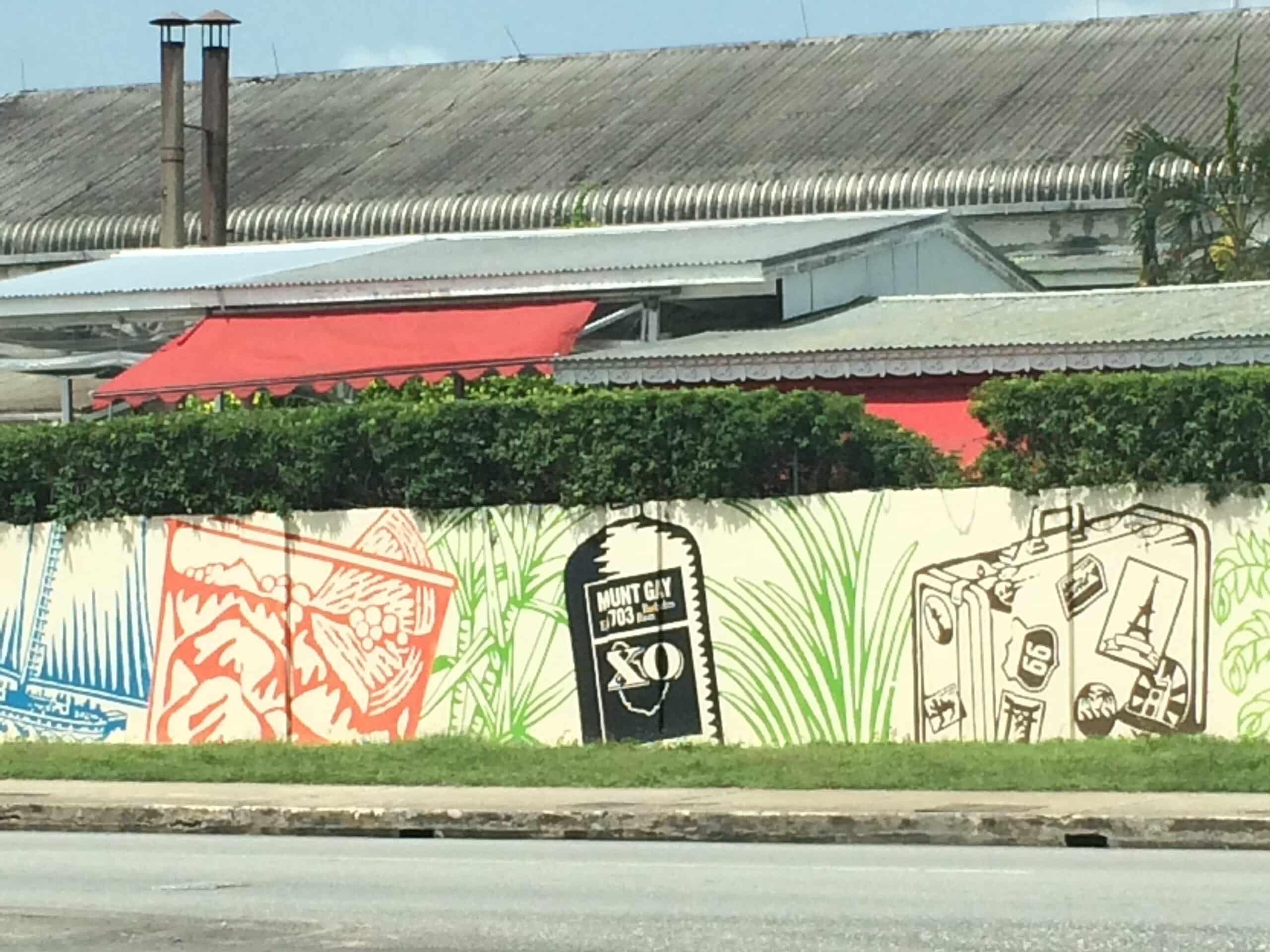 A mural featuring illustrations of a bottle, a hand holding a glass, a Mount Gay XO rum bottle, and a suitcase, painted on a wall beside a corrugated metal industrial building—celebrating the rich tradition of rum in Barbados.
