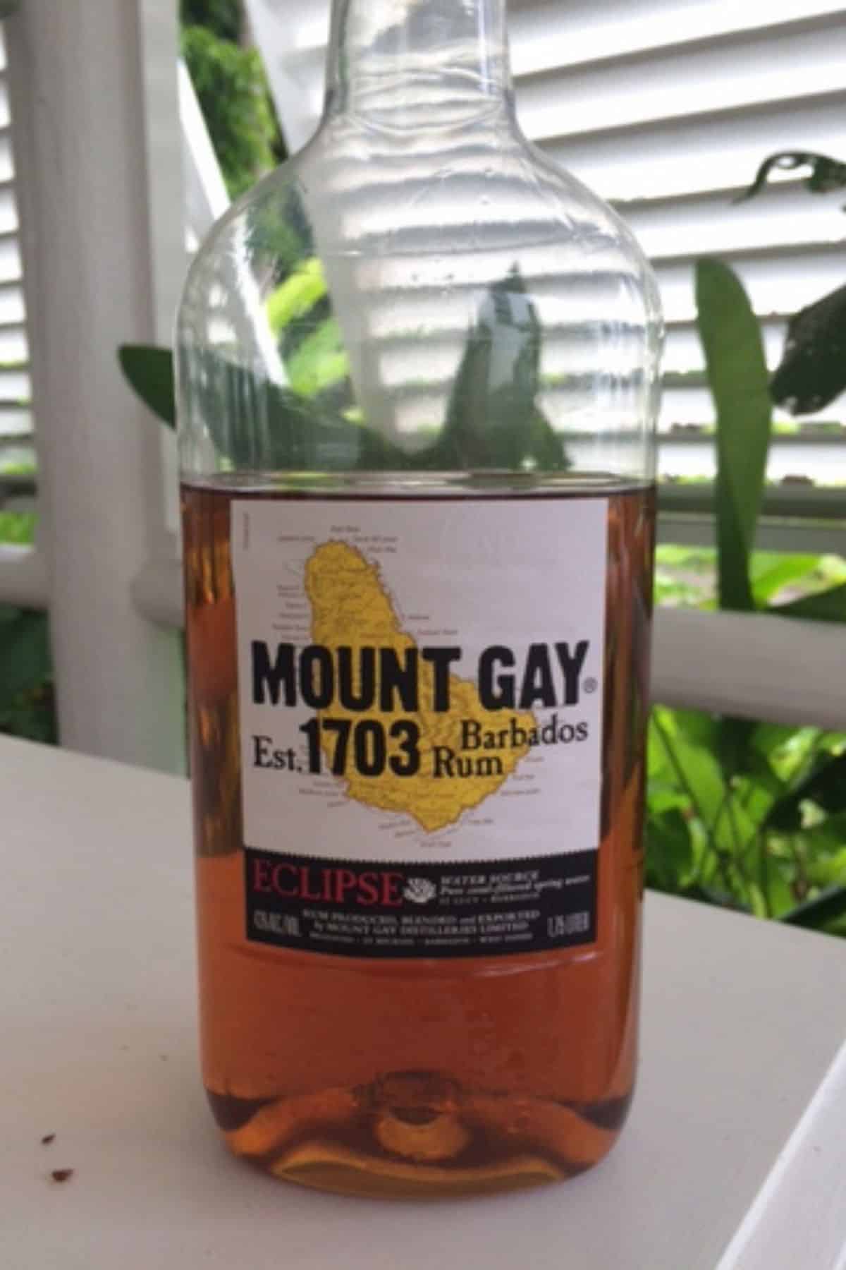 A bottle of Mount Gay Eclipse Barbados Rum, the signature rum in Barbados, with a yellow map label sits on a white surface outdoors, framed by lush plants and a white fence in the background.