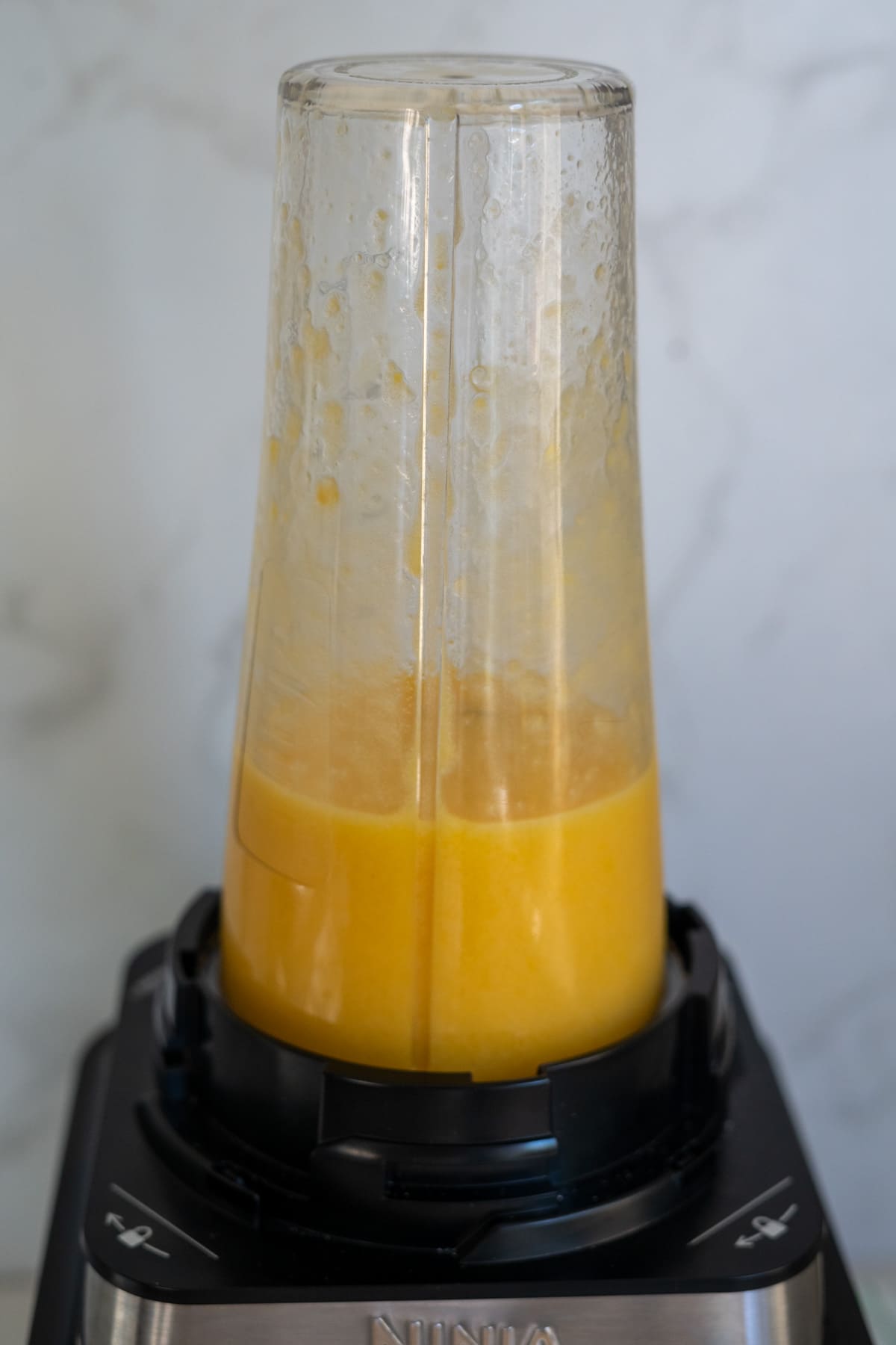 A blender cup filled with yellow-orange liquid is attached upside down to a Ninja blender base, blending the contents.