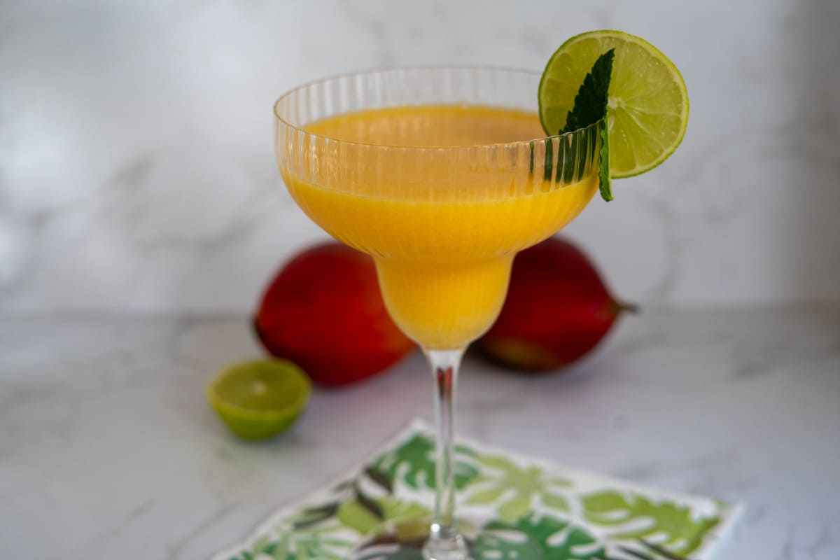 A mango-colored drink in a textured margarita glass, garnished with a lime slice and mint, sits on a leafy napkin with mangoes and a cut lime in the background.