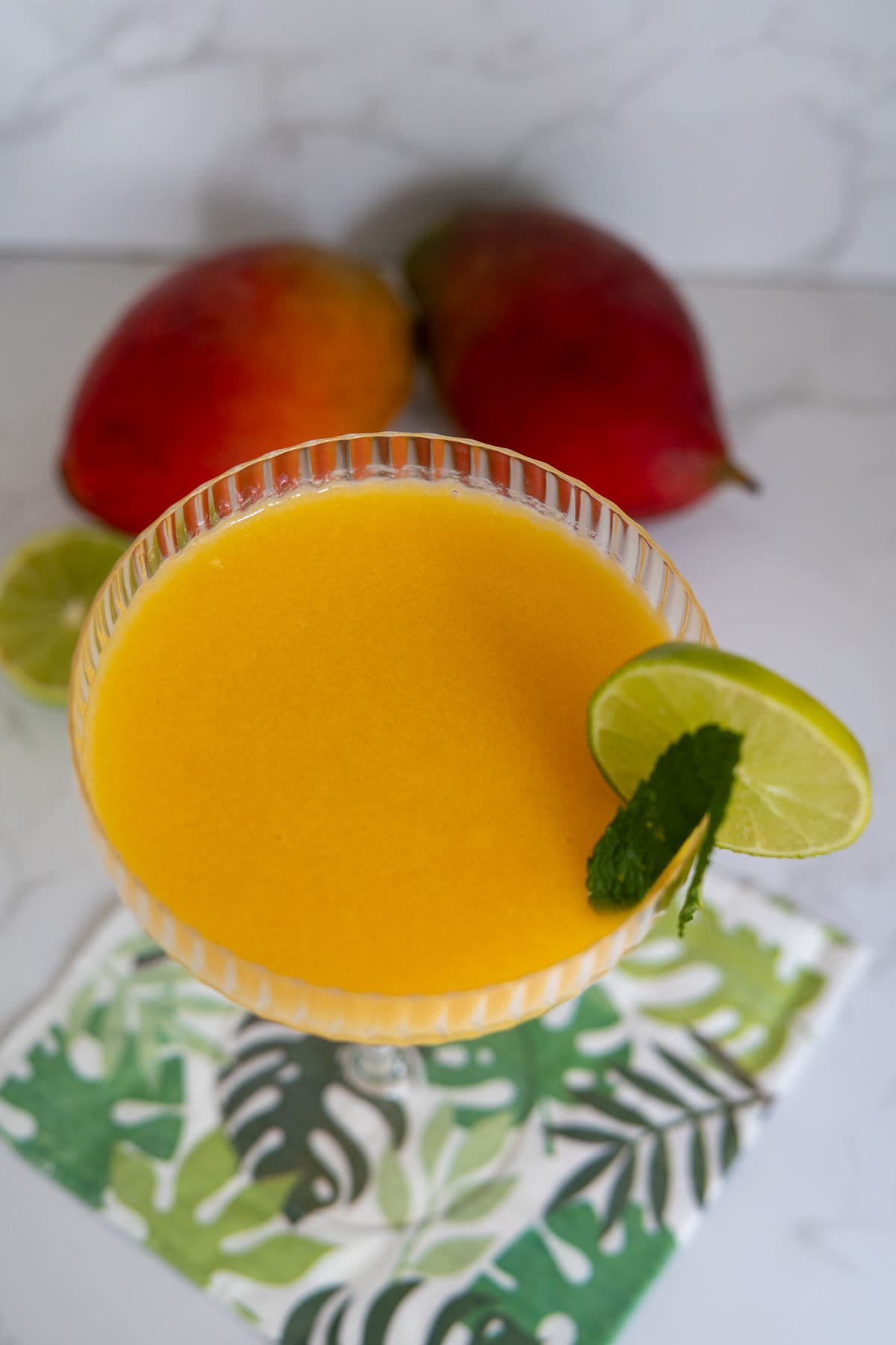 A glass of mango smoothie garnished with a lime slice and mint, placed on a leafy napkin, with two whole mangoes and a lime in the background.