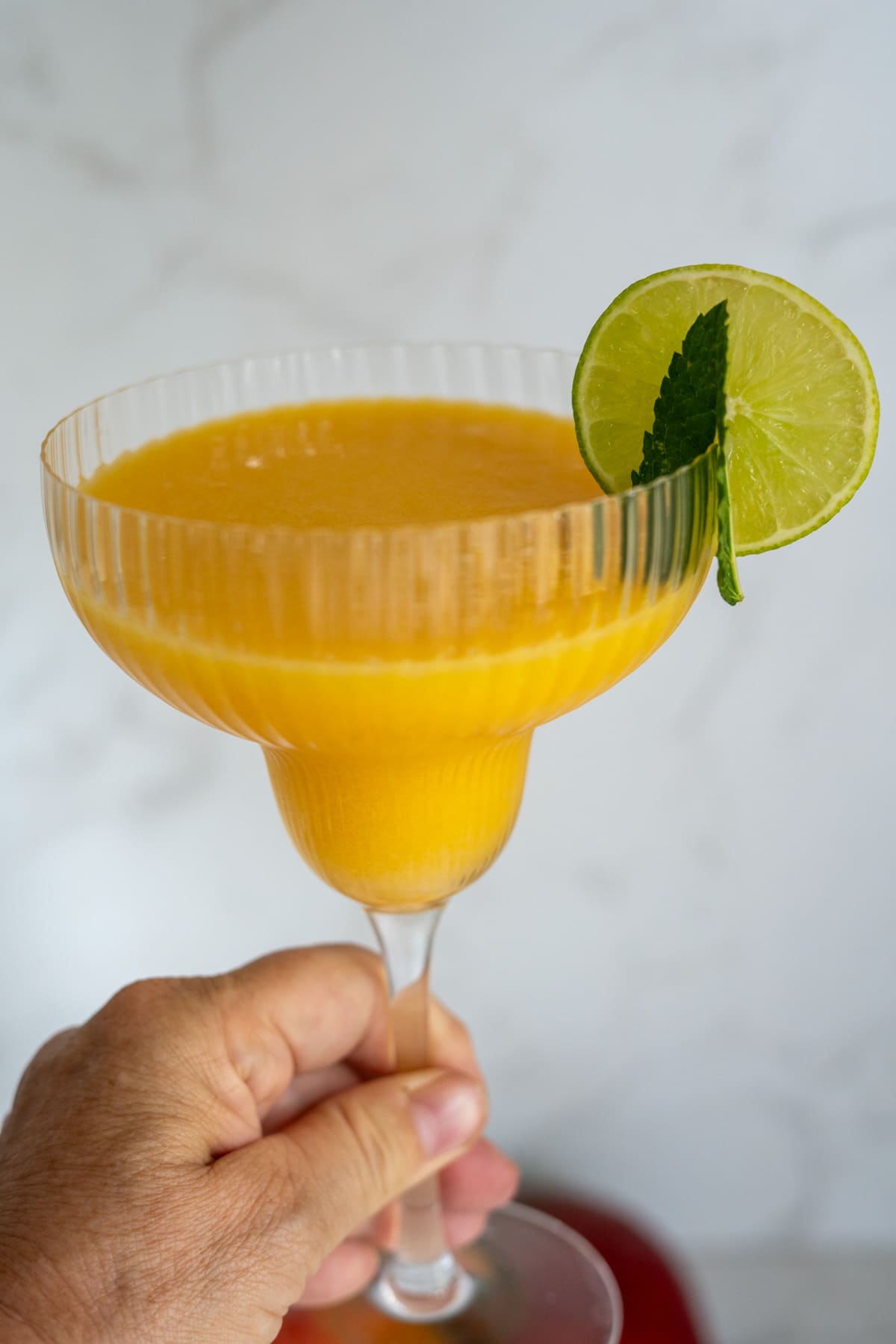 A hand holding a glass of orange-yellow frozen drink garnished with a lime slice and mint leaf against a light background.