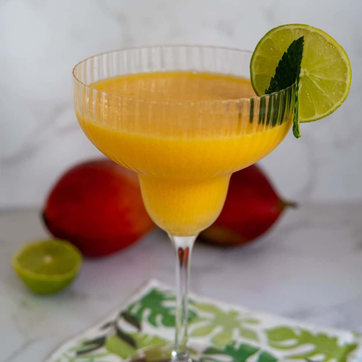 A glass of mango smoothie garnished with a lime wheel and mint leaf, with a mango and half a lime in the background.