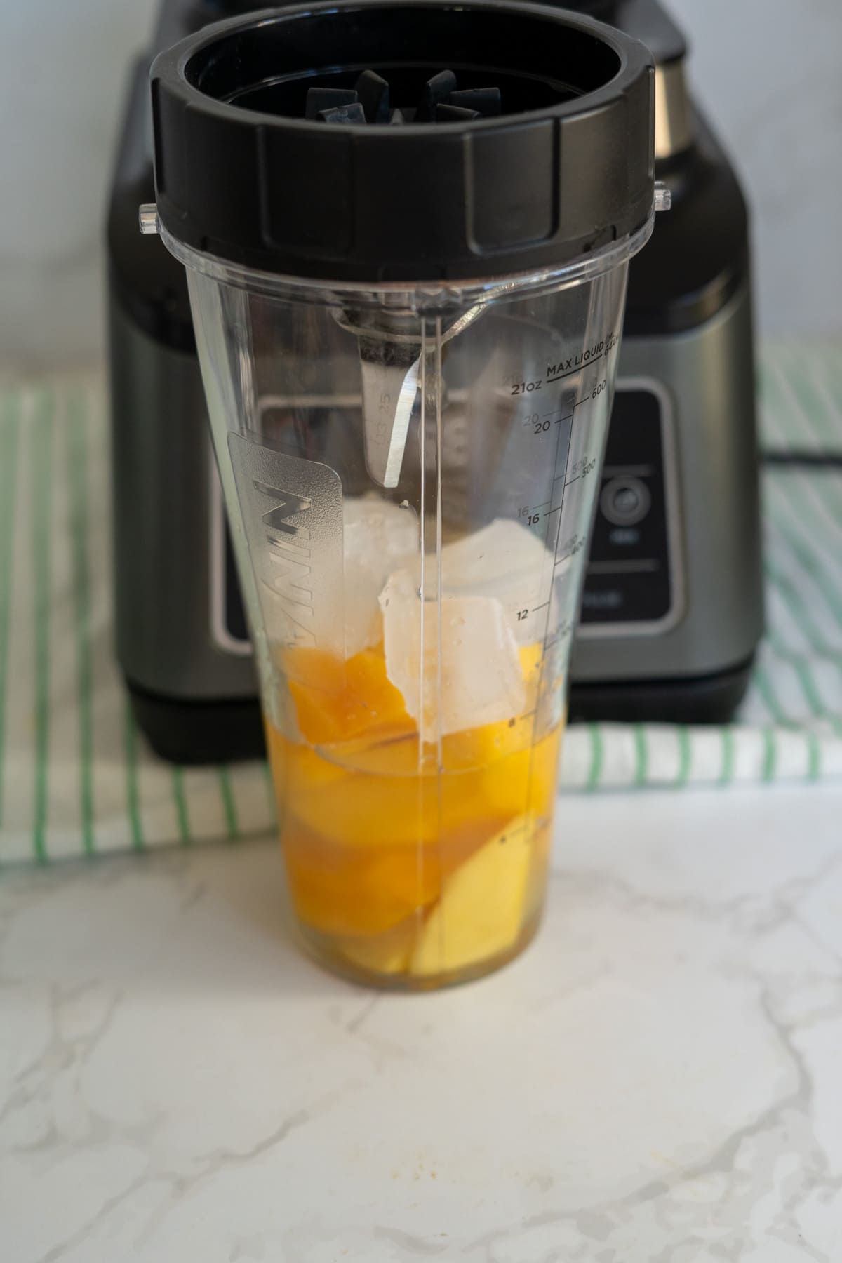 A blender cup with chopped mango, pineapple chunks, and ice cubes placed in front of a blender machine on a striped cloth.